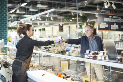 Side view of saleswoman giving package to mature man in organic supermarket