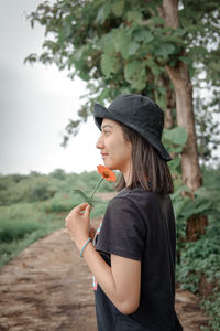 Smiling woman holding flower standing by tree against sky