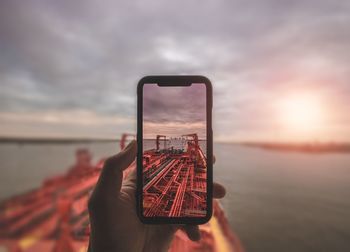 Cropped hand photographing commercial dock at beach against sky