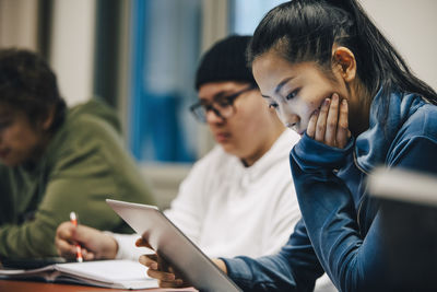 Teenage girl with hand on chin using digital tablet while sitting by classmates in high school