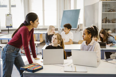 Female teacher talking to schoolgirls sitting with laptops on desk in classroom