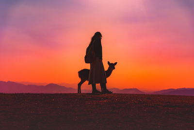 Silhouette woman standing with animal on field against orange sky