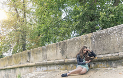 Young woman reading book while sitting by wall