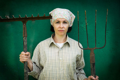 Portrait of woman holding gardening equipment while standing against wall