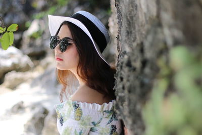 Side view of woman wearing hat and sunglasses while standing by wall