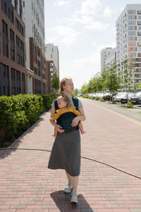 Full length of caucasian mother carrying baby in sling while walking on sidewalk in city