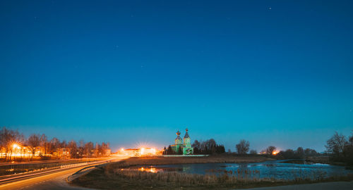 Illuminated road by trees against clear blue sky at night