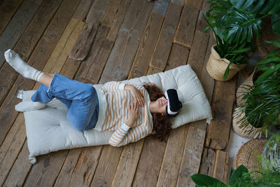 Attractive female wearing virtual reality glasses resting on cozy soft mattress in home greenhouse.