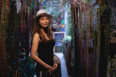 Portrait of smiling woman wearing hat standing amidst creeper plant