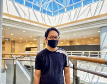 Young man in eyeglasses and face mask standing against architecture and skylight.