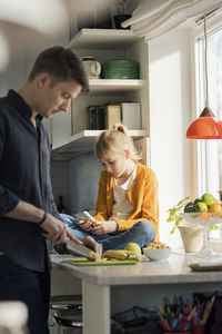 Father slicing fruits white girl using mobile phone at kitchen