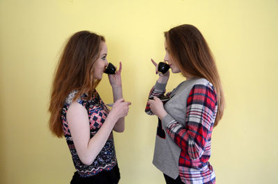 Young women drinking coffee facing each other against yellow wall