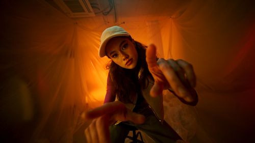 Portrait of young woman with arms raised standing against orange sky