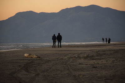 Rear view of silhouette friends walking at beach against mountains during sunset