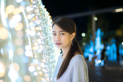 Portrait of young woman standing by illuminated decorative lights at night