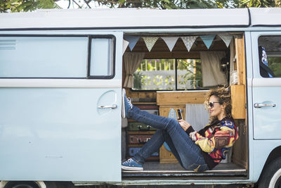 Side view of smiling woman using phone while sitting camper van