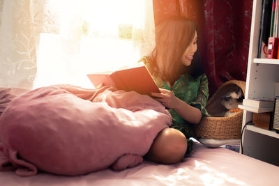 Woman reading book while sitting on bed by cat in basket at home