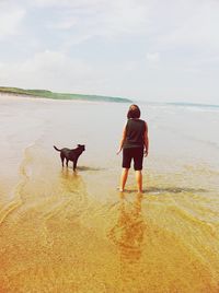 Rear view full length of woman standing with dog at beach against sky