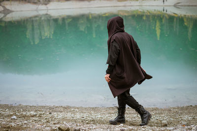 A man in a sweatshirt with a hood walks alone on the beach by a calm lake person