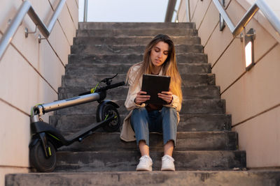 Woman sitting on steps using a tablet with an electric scooter resting on her side. 