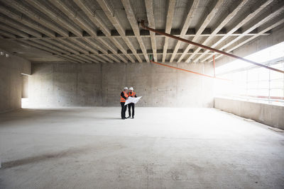 Two men with plan wearing safety vests talking in building under construction