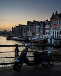 Bicycle by river against buildings in city at sunset