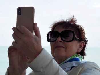 An elderly woman uses a cell phone, takes a selfie from the balcony against the backdrop of the sea.