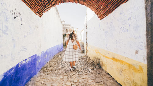  lady in hat and dress going on narrow walkway between old white buildings in city obidos, portugal