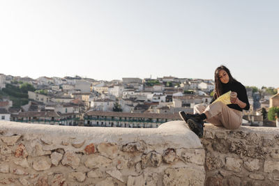 Woman sitting on retaining wall against buildings against clear sky