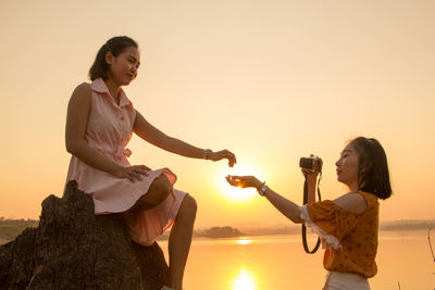 Side view of young woman holding hand while photographing friend against lake and sky during sunset