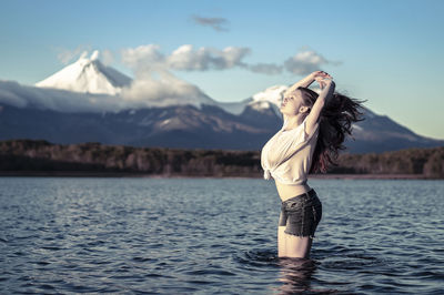 Young woman with arms raised standing in lake against snowcapped mountain