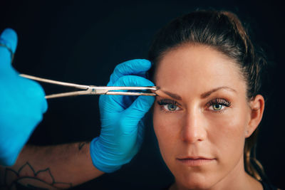 Cropped hands piercing woman eyebrow against black background