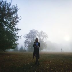 Full length of young woman standing on grassy field against sky during foggy weather