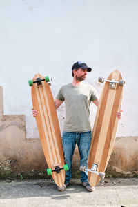 Bearded male skateboarder in cap and t shirt looking away while standing on walkway with handmade longboards