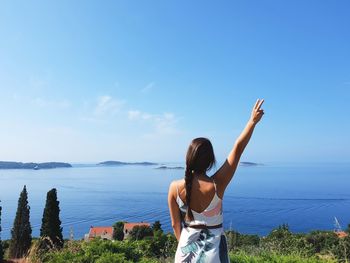 Rear view of woman with hand raised standing by sea against blue sky during sunny day