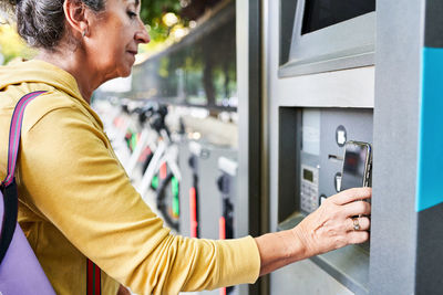 Crop senior female using smartphone to gain access to atm near electric bike rental station on city street