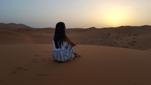 Woman sitting on sand dune at desert against sky