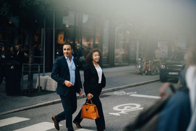 Full length of smiling business coworkers talking while crossing street in city