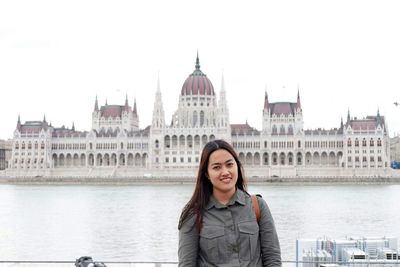 Portrait of smiling woman standing against river and hungarian parliament building in city