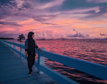 Woman standing on railing against sea during sunset