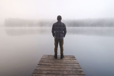 Rear view of man standing on pier over lake during foggy weather