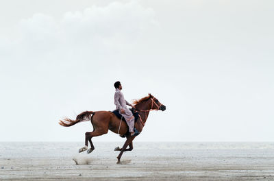 Side view full length of man riding horse on sand against clear sky