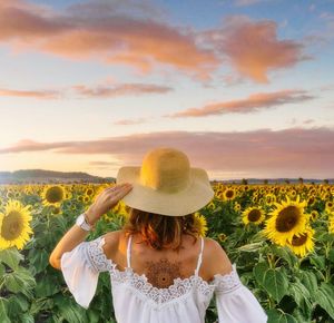 Rear view of young woman standing on sunflower field against cloudy sky during sunset