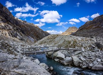 Footbridge over stream by mountains against cloudy sky