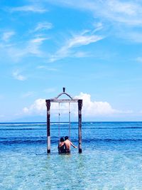 Rear view of couple sitting on swing in sea against blue sky