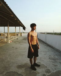 Full length of shirtless man standing on building terrace against clear sky