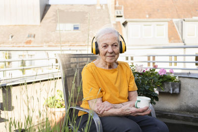 Smiling senior woman holding coffee cup listening music through wireless headphones on balcony