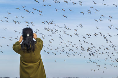 Low angle view of woman standing against flying birds in sky