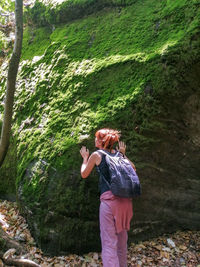 Full length of woman standing by tree in forest, touching and smelling moss