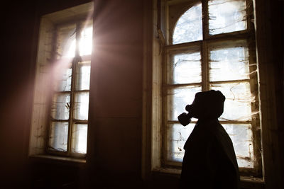Silhouette woman wearing gas mask standing by window in abandoned building